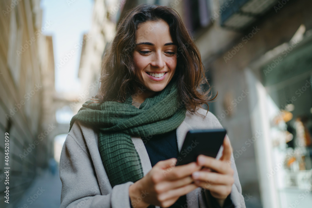 © Roman - Portrait of young smiling dark-haired woman wearing light-colored coat and green scarf, walking city streets and using smartphone to interact with her friends. Good vibes. Modern technologies concept © Roman - Portrait of young smiling dark-haired woman wearing light-colored coat and green scarf, walking city streets and using smartphone to interact with her friends. Good vibes. Modern technologies concept