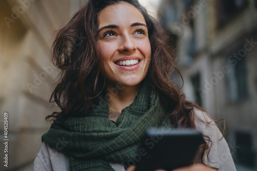 Young attractive dark-haired Caucasian woman walking streets, holding smartphone and looking away. Good vibes and positive emotions. Concept of online communication using mobile digital technologies photo