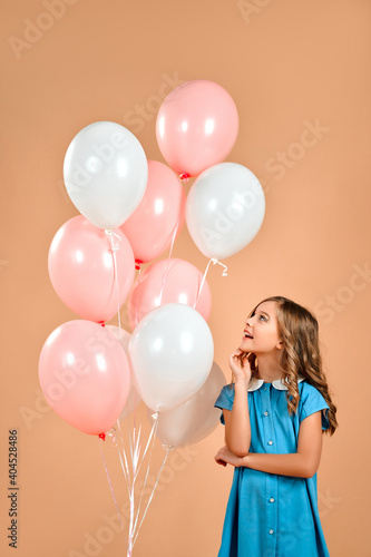 A cute girl in a blue dress and curly hair holds pink and white balloons and smiles cheerfully isolated on a peach gentle background.Valentine's Day. © Valerii Apetroaiei