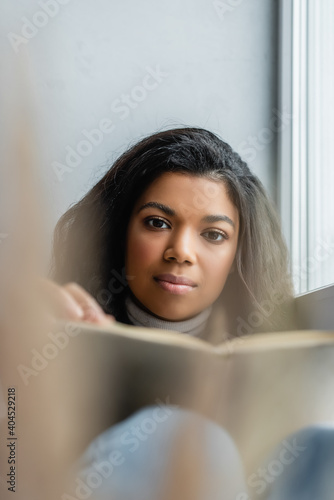 young african american woman looking at camera while holding book at home, blurred foreground