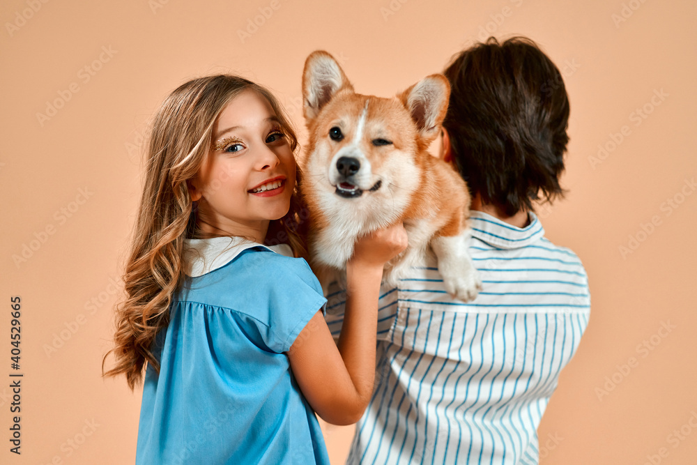 A boy stands with his back to the camera and a beautiful girl with curls in a dress and holds a cute funny dog ​​on his shoulder isolated on a powdery peach background.Valentine's Day.