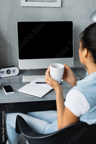 young african american freelancer holding cup of tea near computer monitor, smartphone and notebook on desk, blurred foregroud photo