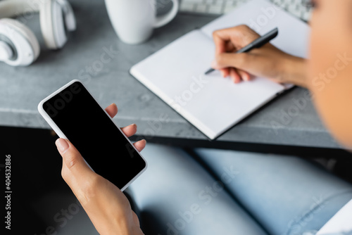 partial view of african american woman holding smartphone with blank screen and writing in notebook, blurred foreground