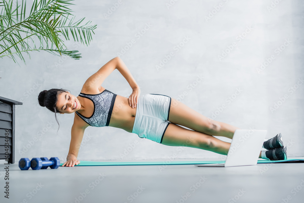Fototapeta premium happy african american sportswoman exercising in side plank pose on fitness mat near laptop and dumbbells