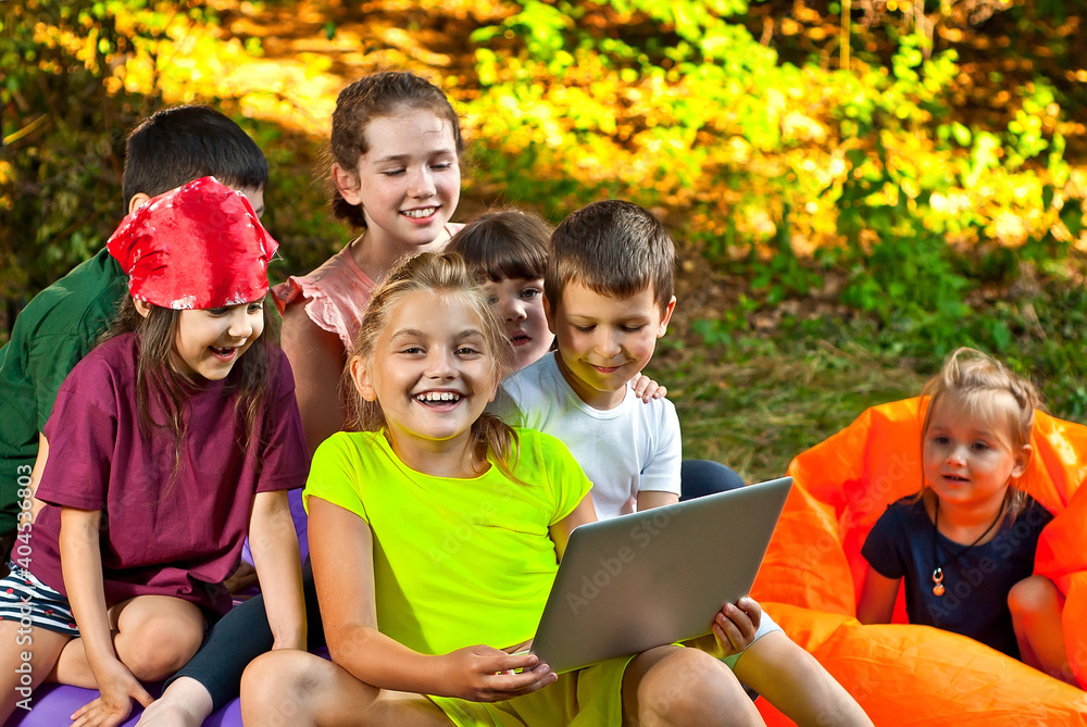 Children play in nature with a laptop. Boys and girls are resting in a tent camp. Children sit on the carpet on the grass in summer. Children play in nature with a laptop. Boys and girls are resting in a tent camp. Children sit on the carpet on the grass in summer.