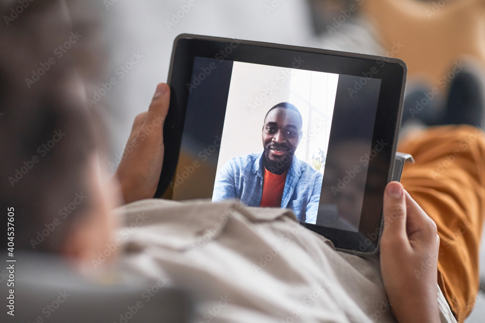 Unrecognizable African American kid relaxing on sofa holding digital tablet in his hands having online call with his dad