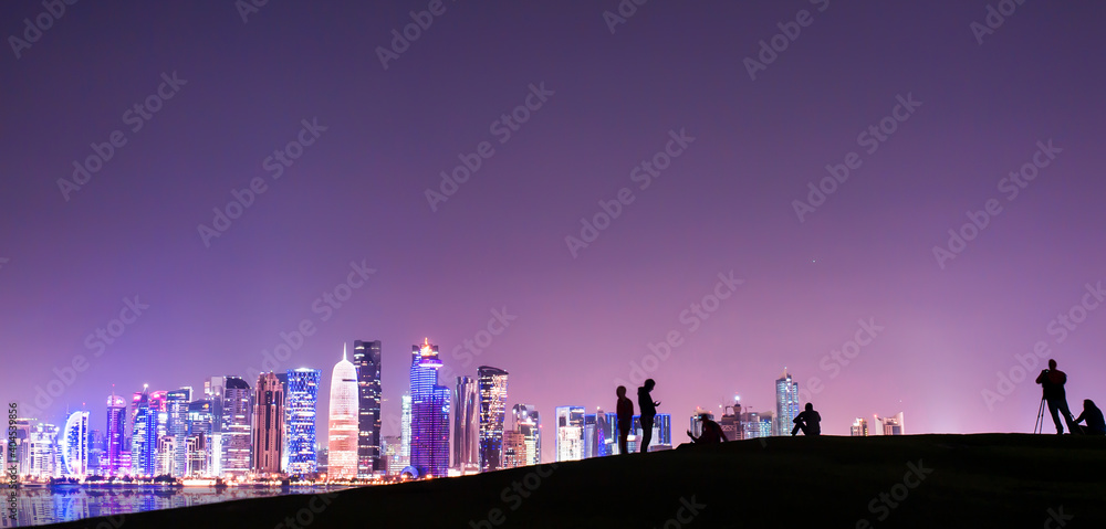 (Selective focus) Stunning panoramic view of the Doha skyline illuminated at dusk in the distance and some people relaxing on a hill in the foreground.