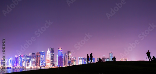 Obraz na płótnie (Selective focus) Stunning panoramic view of the Doha skyline illuminated at dusk in the distance and some people relaxing on a hill in the foreground. w salonie