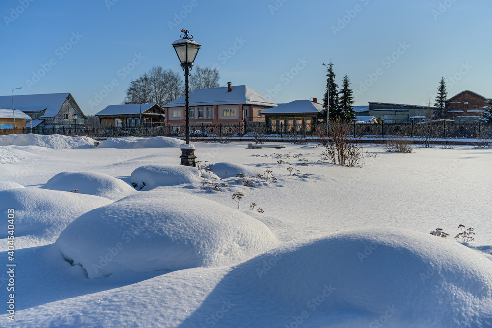 in the park of the city of Nevyansk (Russia) in winter. a lot of white fluffy snow on the ground and plants, snow balls formed. in the distance a lantern, trees and houses. blue sky