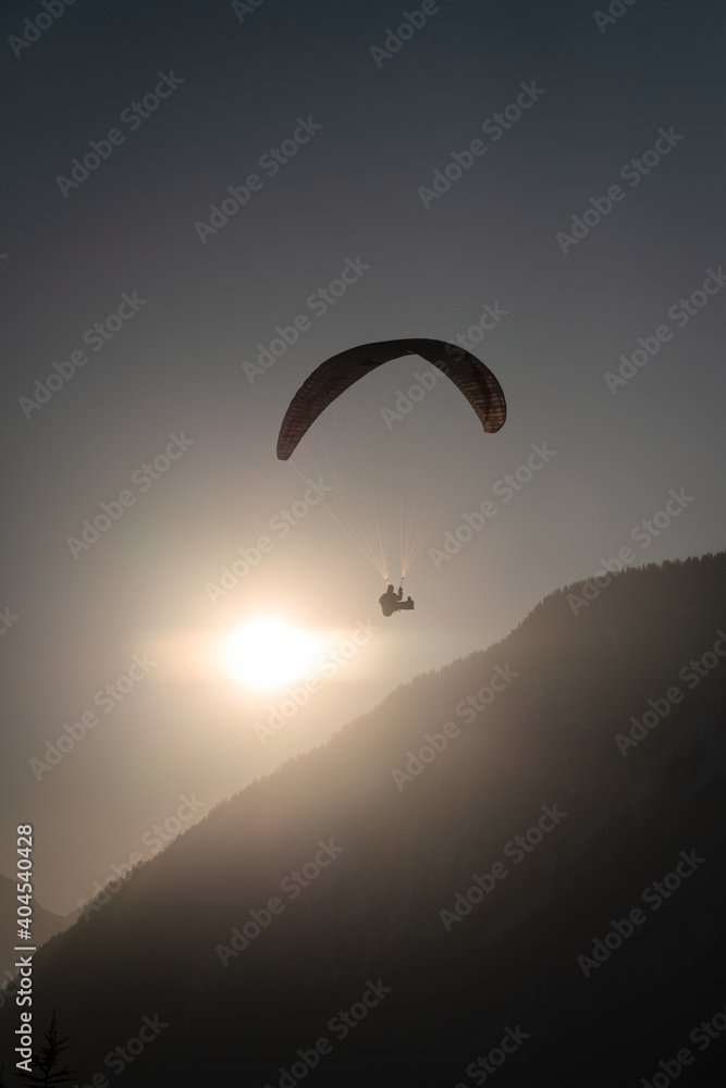 Paraglider over the silhouette of a wooded mountain ridge with the sun backlit at sunset Paraglider over the silhouette of a wooded mountain ridge with the sun backlit at sunset