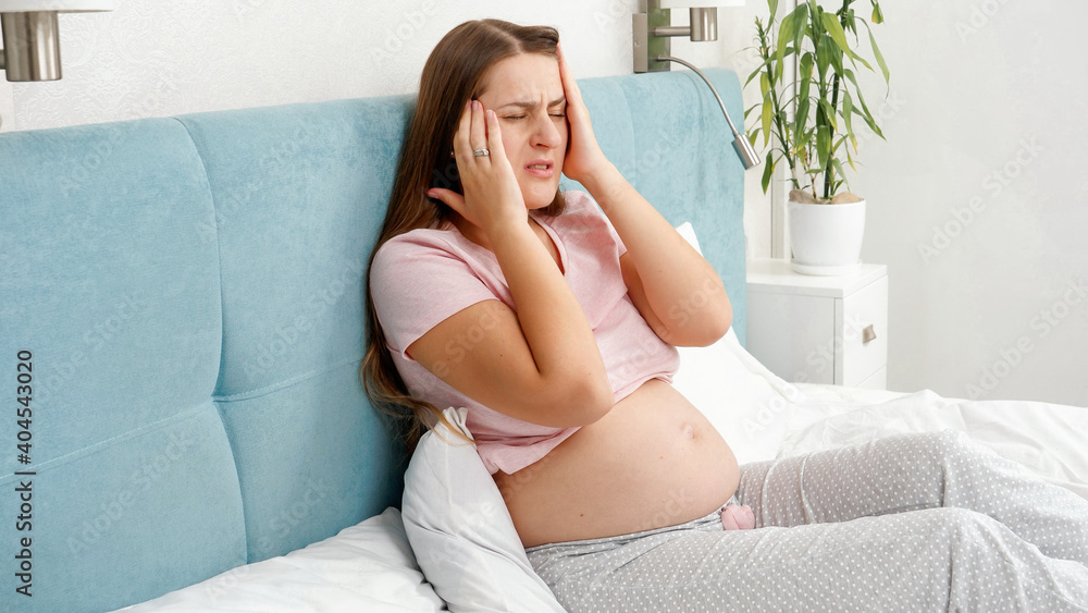 Portrait of young pregnant woman suffering from headache lying in bed. Concept of healthcare and pain during pregnancy Portrait of young pregnant woman suffering from headache lying in bed. Concept of healthcare and pain during pregnancy