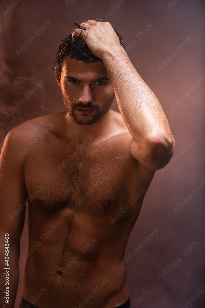sexy, wet man with muscular torso posing with hand on head on dark background with smoke sexy, wet man with muscular torso posing with hand on head on dark background with smoke