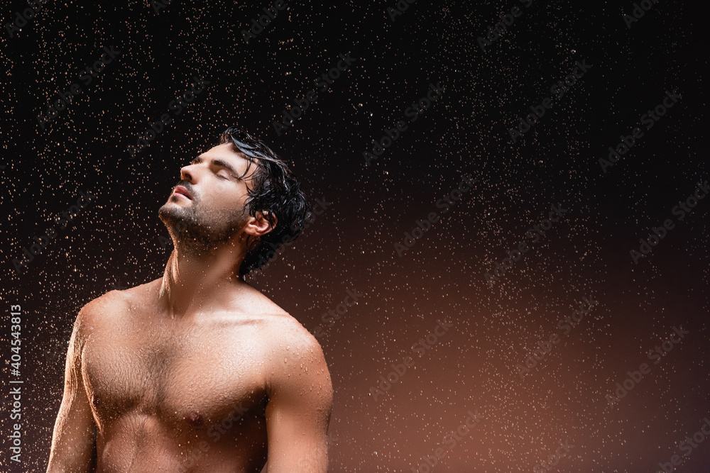 shirtless man with muscular torso standing on dark background under falling rain