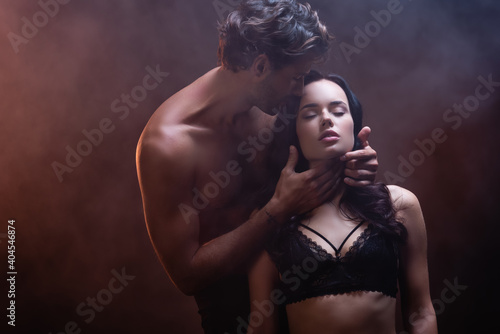 shirtless muscular man hugging neck of seductive woman in black lace bra on dark background with smoke