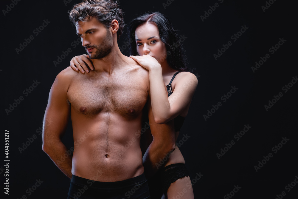 sensual brunette woman embracing shoulders of man with muscular torso while looking at camera isolated on black