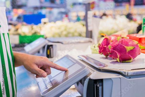 The seller is weighing fruit and points the fingers on the screen electronic scales in supermarket.