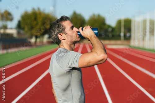 Mature fitness man on red running track, drinking from blue bottle. Copy space. Copy space. Close up. Lifestyle and sports