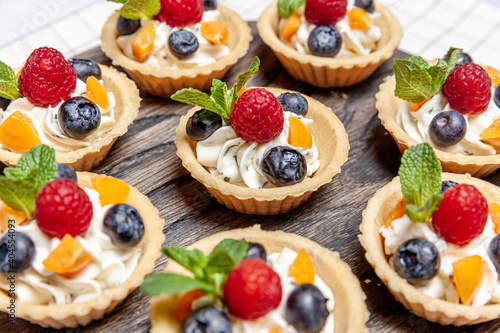 Fruit and berry tartlets dessert assorted on wooden tray. Closeup of delicious pastry sweets pies colorful cakes with fresh natural raspberry blueberry and cheese cream. French bakery catering.