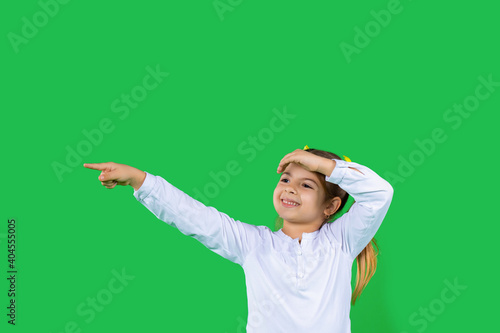 A cute little girl smiles, looks into the distance and shows her index finger. Green background. Empty space.