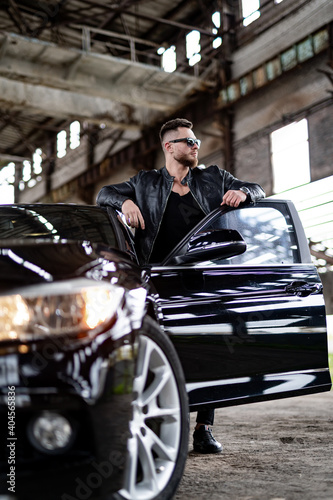 Handsome fashion model guy near opened door of luxury car. Total black outfit on man near dark car. © Vadim