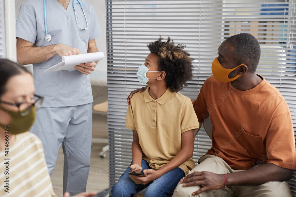 Fototapeta premium Portrait of African-American family wearing masks and looking at doctor while waiting in line at medical clinic
