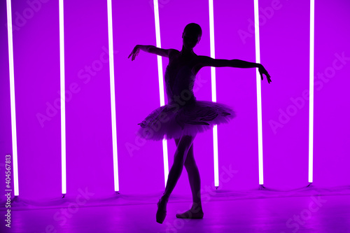 Young classical ballet dancer posing in a dark studio against a background of purple neon lights. Slender ballerina is silhouetted on tiptoes in white tutu. Ballet school poster.