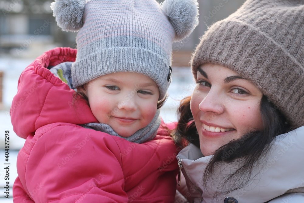 Little girl in a knitted hat with buboes in the arms of a smiling mother on the street in winter Little girl in a knitted hat with buboes in the arms of a smiling mother on the street in winter