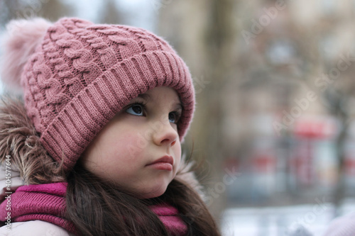 Little smiling white girl with long dark hair in a knitted hat on a background of snow and blurred buildings © PeterPike