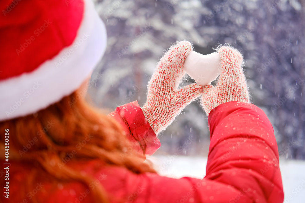 A woman in a santa hat holds a heart made of snow against a background of forest trees, close-up. Woman holding a heart shaped snowball for valentine day