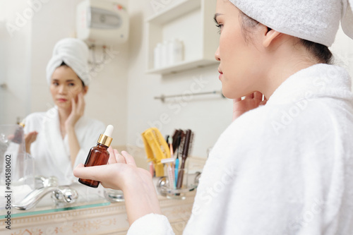 Young woman in bathrobe and towel on head applying serum when standing at mirror in bathroom