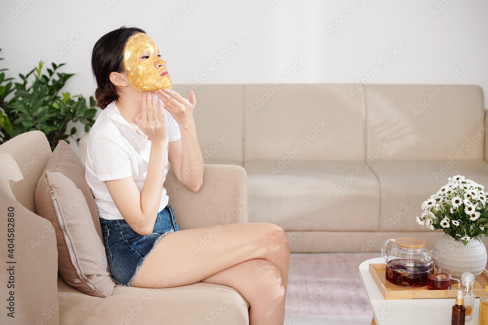 Pretty young Asian woman in denim shorts and t-shirt sitting on sofa and applying silicone sheet mask on face Pretty young Asian woman in denim shorts and t-shirt sitting on sofa and applying silicone sheet mask on face