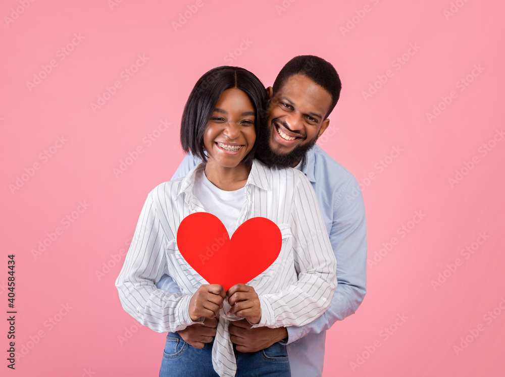 Valentine's Day holiday. Portrait of positive black couple holding red paper heart and hugging on pink background Valentine's Day holiday. Portrait of positive black couple holding red paper heart and hugging on pink background