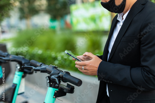 Electric scooter and mobile phone. Businessman in medical mask using e vehicle rent service with smartphone in urban city street and park. 