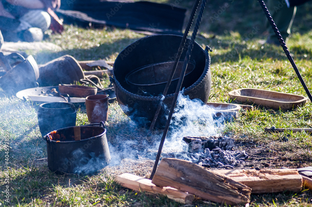 Homemade food in iron pot, a cauldron in smoke near dying embers, historical reenactment of Slavic or Viking lifestyle. - obrazy, fototapety, plakaty 