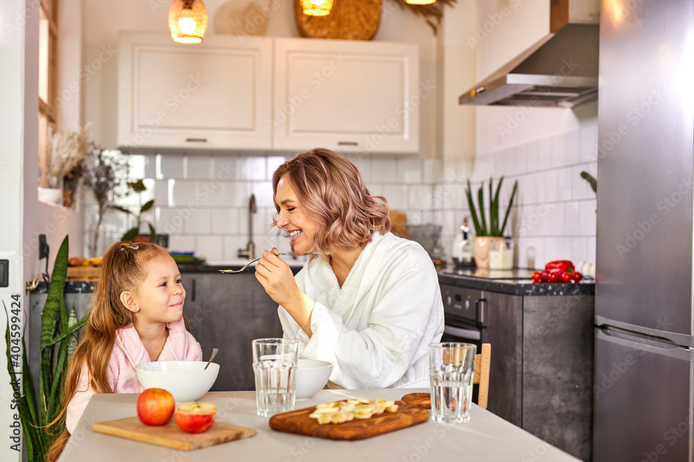 mother feeding her daughter by spoon, eating porridge, fresh fruits in the kitchen