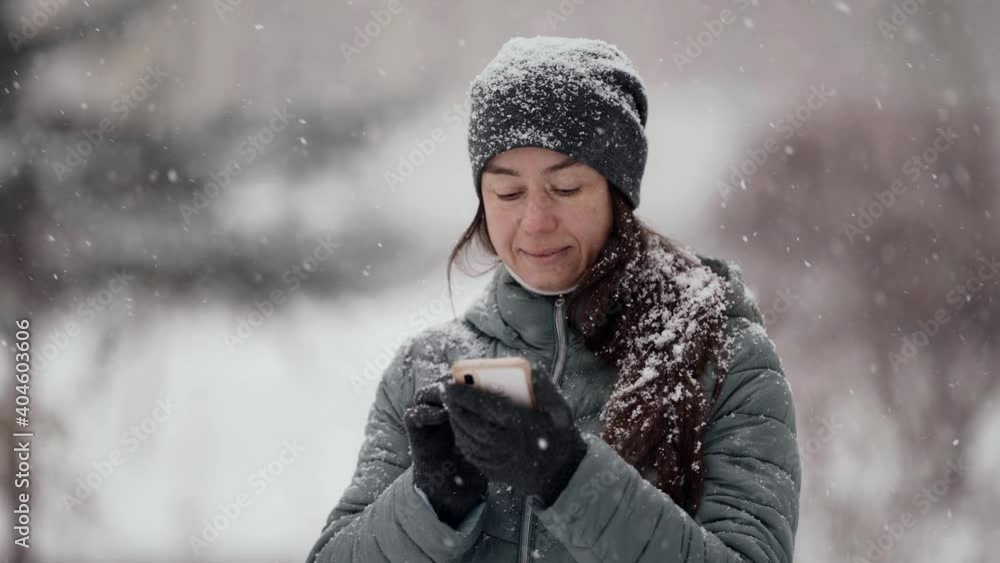 Portrait of a dark-haired middle-aged woman in a hat and jacket, she is outside on a winter day, she holds a phone in her hands. Snowy weather in the city park. Snow is falling. Portrait of a dark-haired middle-aged woman in a hat and jacket, she is outside on a winter day, she holds a phone in her hands. Snowy weather in the city park. Snow is falling.