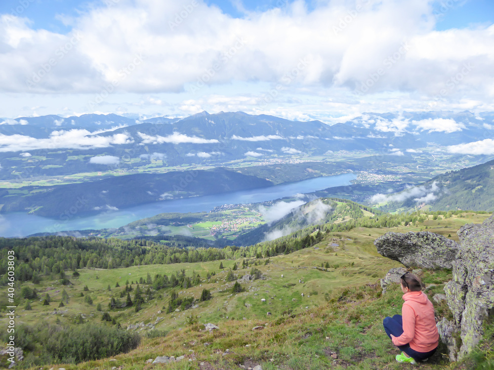 Naklejka premium A woman in orange hoodie squatting under a rock and enjoying the view on the Millstaetter lake from Granattor in Austrian Alps. The distant lake is surrounded by high mountains. Few clouds above.