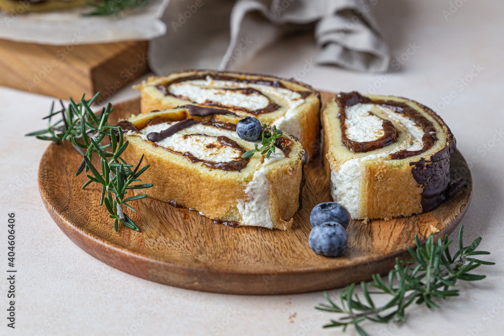Sponge cake roll with chocolate and cream cheese decorated with chocolate glaze, blueberry and rosemary on parchment, light concrete background. Biscuit swiss roll. Sponge cake roll with chocolate and cream cheese decorated with chocolate glaze, blueberry and rosemary on parchment, light concrete background. Biscuit swiss roll.
