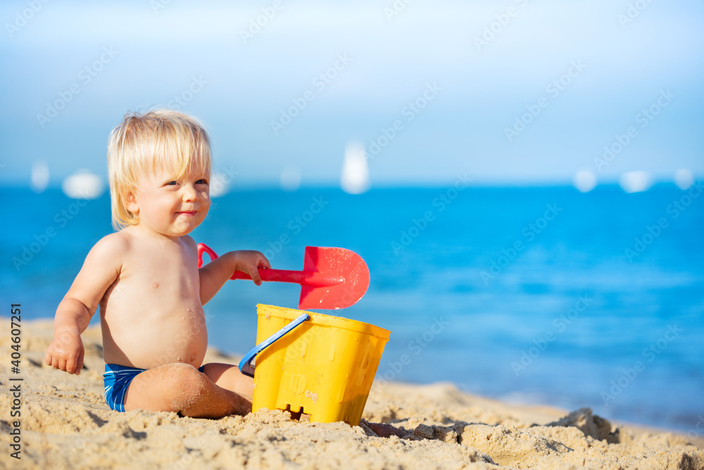 Little happy blond boy play with small bucket on the beach near the sea Little happy blond boy play with small bucket on the beach near the sea
