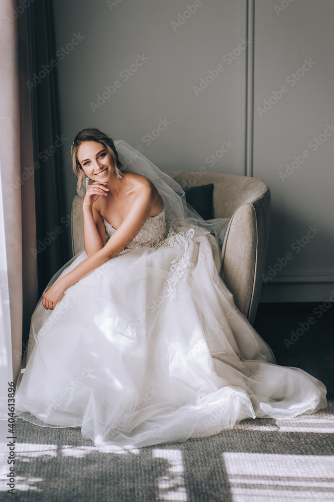 A beautiful, sweet, smiling blonde bride in a white lace dress with a long veil sits on an armchair against the background of a wall in a hotel room. Wedding model portrait. A beautiful, sweet, smiling blonde bride in a white lace dress with a long veil sits on an armchair against the background of a wall in a hotel room. Wedding model portrait.