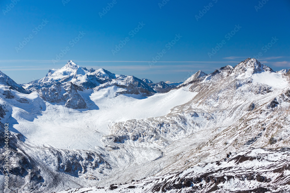 Obraz premium Snow-capped peaks and clear blue skies. Winter mountains, alpine skiing. Sunny snowy landscape. Panoramic mountain view. Rocks and white slopes covered with glaciers