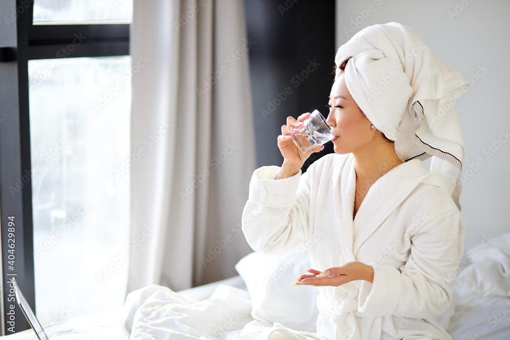 chinese woman taking a pill, sitting on her bed at home, wearing bathrobe and towel, holding medicaments or vitamins and glass of water chinese woman taking a pill, sitting on her bed at home, wearing bathrobe and towel, holding medicaments or vitamins and glass of water