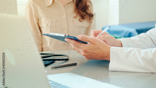 Close-Up Hands of Male Doctor Fills Out a Patient Card, Young Woman Patient Sitting near on the Background. Talking With a Doctor at Consultation During an Appointment.