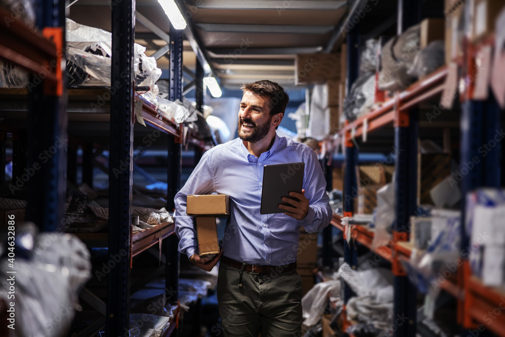 Fototapeta premium Smiling bearded businessman walking trough storage of shipping firm, holding boxes under armpit and using tablet to check on goods.