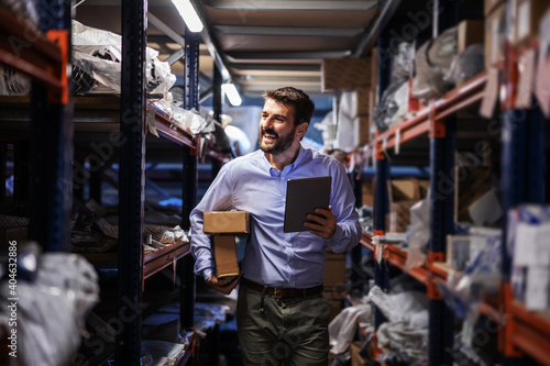 Smiling bearded businessman walking trough storage of shipping firm, holding boxes under armpit and using tablet to check on goods.