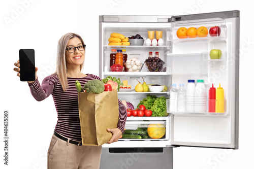 Young woman in front of a fridge holding a grocery bag and showing a smartphone
