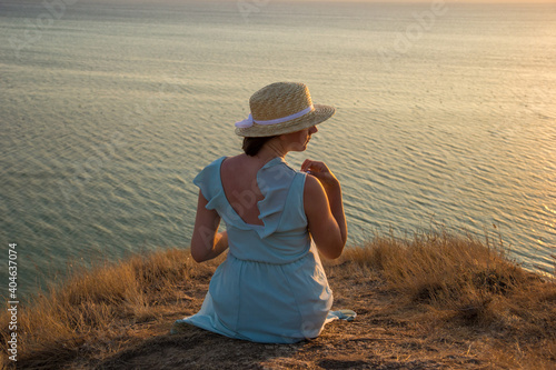 A pretty girl in a long blue dress and straw hat sits on a high cliff by the sea. Young beautiful woman in the hat with white ribbon watching sunset on the shores of the calm sea.