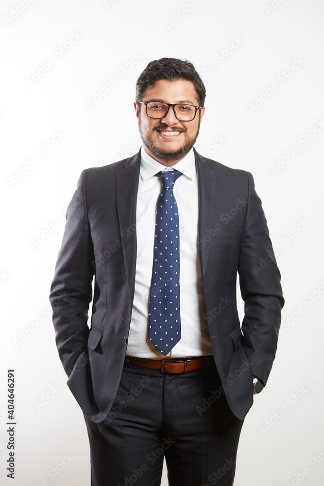 A young Latino businessman wearing glasses and standing in a dark suit and tie on a white background A young Latino businessman wearing glasses and standing in a dark suit and tie on a white background