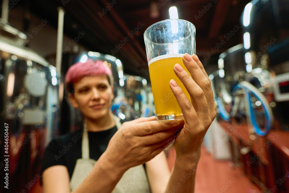 Hand of female brewer in apron examining glass of fresh beer after preparation - obrazy, fototapety, plakaty 