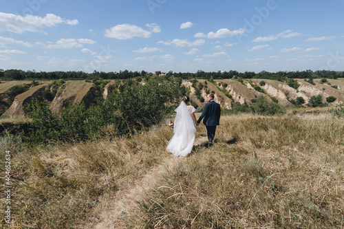 Stylish groom in a blue suit and a beautiful curly-haired bride in a white dress are walking in nature, against the background of the hills, holding hands. Wedding portrait of newlyweds in love.